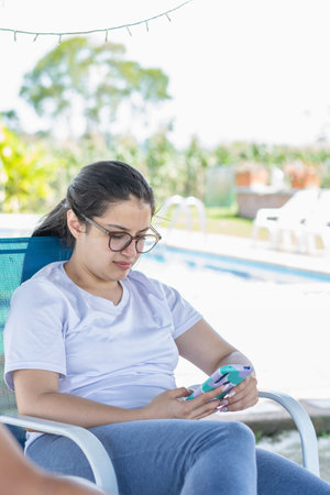 young latina woman with glasses, sitting on a chair, looking at social networks on her cell phone.の写真素材