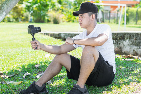 Young Latino man sitting on the grass, recording himself with a camera, making a vlog of his tripの写真素材