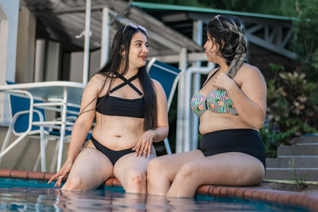 latina friends sitting by the pool with their feet in the water talking and gossiping about their lives.の写真素材