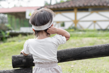 View of the back of a little brunette girl standing next to the wooden fence, touching her head while looking at natureの写真素材