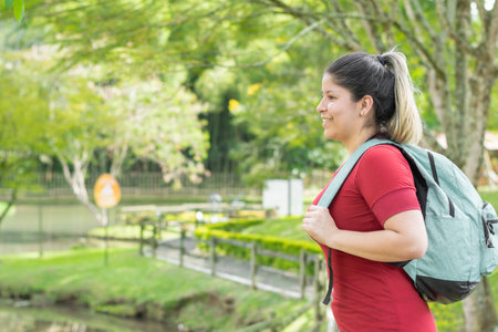 beautiful young latina college student woman, carrying a big bag while smiling and taking a walk in the parkの写真素材