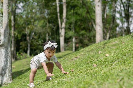 little brown girl trying to climb up the side of a small hillside covered with green grassの写真素材