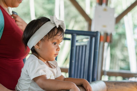 Beautiful brunette Latina baby looking attentively while her mother holds her next to the wooden fenceの写真素材