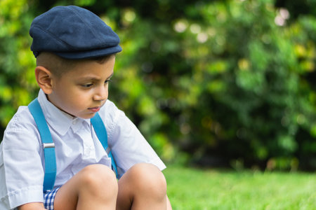very sad little latino boy sitting on the ground in a public park looking down at the groundの写真素材
