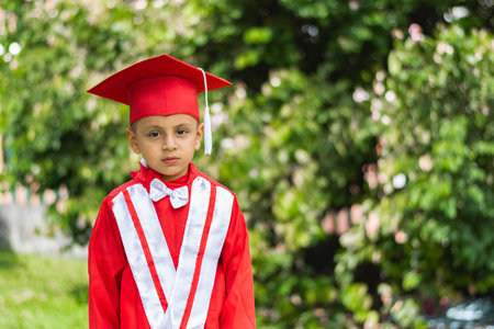 portrait of bored little latin boy, dressed in red graduation gownの写真素材