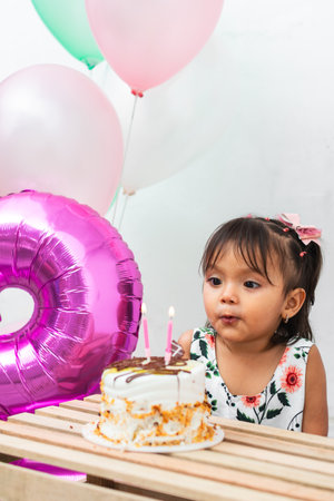 little latin brunette girl in front of her birthday cake, blowing out the two candles to blow them...
