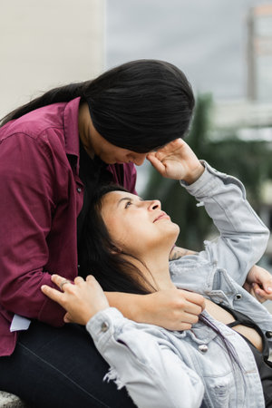 young latina lesbian woman lying on her partner's lap looking up at her while caressing his faceの写真素材