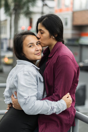 young latina woman looking sideways while her girlfriend kisses her with her eyes closed, lesbian couple in love and happyの写真素材