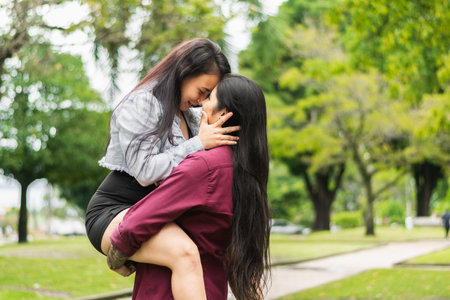 young latin lesbian woman holding her partner in her arms while they put their faces together and look at each other very much in love, in the background of a green park.の写真素材