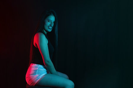 young latin woman sitting sideways looking and smiling, illuminated with red and aquamarine green studio lights.の写真素材