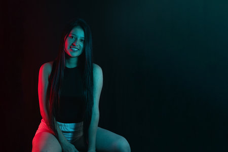 Young latin woman posing under red and teal lights, beautiful girl smiling and looking straight at the cameraの写真素材