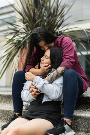young latina lesbian couple sitting on the street, kissing their girlfriend on the forehead while hugging her from behind.の写真素材