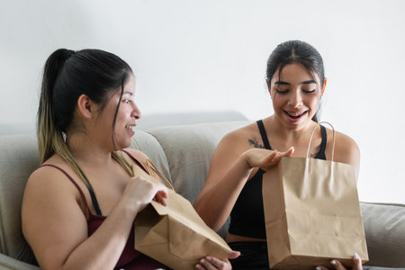 young latina woman very surprised looking at what is inside the shopping bag, next to her friend sitting on the couchの写真素材