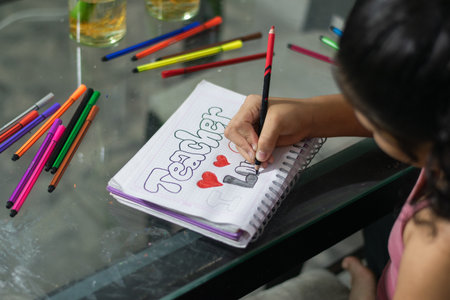 Close-up side view of girl's hand drawing colorful letters in a notebook on a glass table