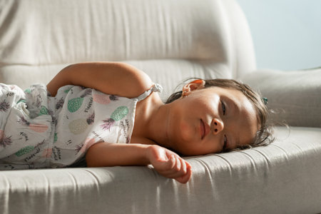 Little girl sleeping on a light gray sofa, afternoon light, arm relaxed over the edge, peaceful nap at home.の写真素材