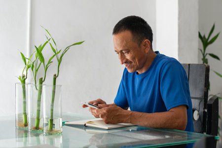 Smiling Latino man at a table using a smartphone, informal home scene, with four fingers amputated on his left hand.の写真素材