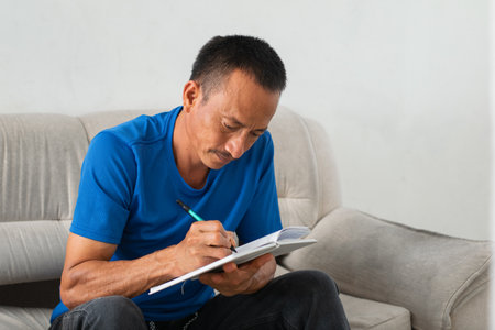 Focused Latino man writing in notebook on his lap, seated on couch, left hand missing four fingers evidentの写真素材