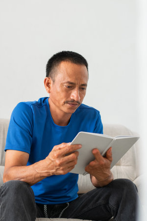 Man in a blue shirt reading his white diary while sitting on a sofa, focused, his left hand showing the amputation of his fingers.の写真素材
