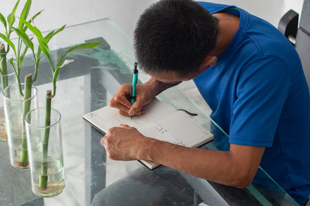 Top view of a man leaning over a glass table writing in a notebook, focused, with visible amputation of his left fingers.の写真素材