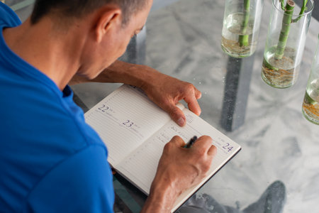 Over shoulder view of man writing in open planner at table, concentrated, left hand missing four fingers clearの写真素材