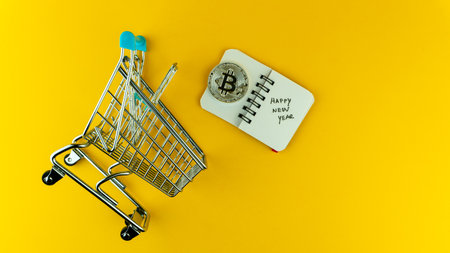 Overshot of a small shopping cart, bitcoin and a small notebook with Happy New Year text on it in yellow background. Business planning concept.の写真素材