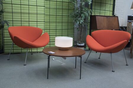 Two orange chairs with fabric covered with gray iron legs, with a table in front of them with a black iron frame with wooden tabletop. With a light green back wall and a gray concrete floorの写真素材