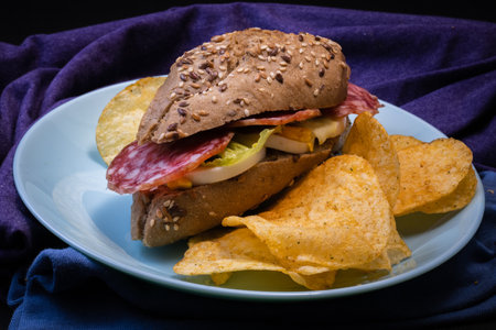 Sandwich with a rustic bun,salami,chips,salad,pickles,onion and a boiled egg on a blue plate