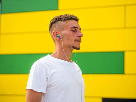 A Caucasian male using his phone on yellow wall background and listening music with headphones