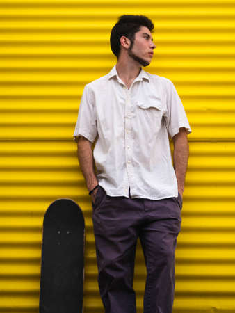 A vertical shot of a young Caucasian man with his skateboard near a yellow wallの写真素材