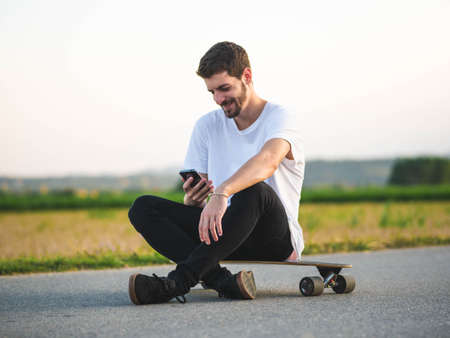 A handsome Caucasian male sitting on a longboard and using a phone at outdoorsの写真素材
