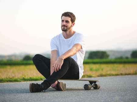 A handsome Caucasian male sitting on a longboard and using a phone at outdoorsの写真素材