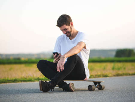 A handsome Caucasian male sitting on a longboard and using a phone at outdoorsの写真素材