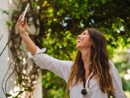 beautiful attractive female taking a selfie with her smartphone. Tourist taking selfie in a street surrounded by trees. Woman taking selfie standing at the beachの写真素材