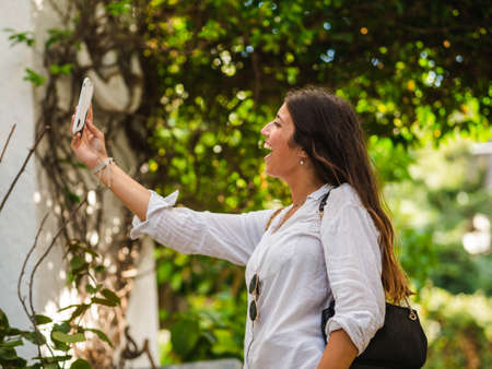 beautiful attractive female taking a selfie with her smartphone. Tourist taking selfie in a street surrounded by trees. Woman taking selfie standing at the beachの写真素材