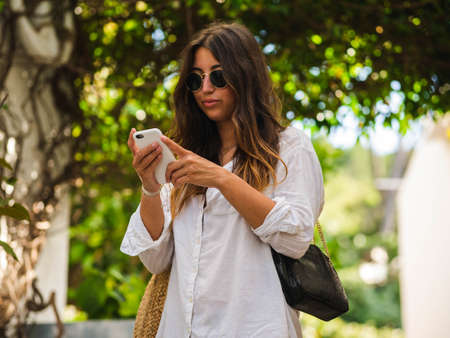 beautiful attractive female taking a selfie with her smartphone. Tourist taking selfie in a street surrounded by trees. Woman taking selfie standing at the beachの写真素材