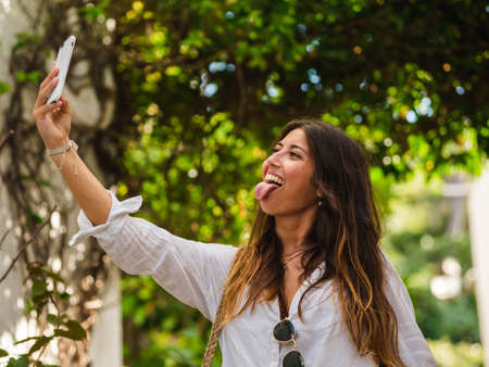 beautiful attractive female taking a selfie with her smartphone. Tourist taking selfie in a street surrounded by trees. Woman taking selfie standing at the beachの写真素材