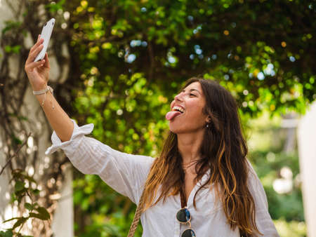 beautiful attractive female taking a selfie with her smartphone. Tourist taking selfie in a street surrounded by trees. Woman taking selfie standing at the beachの写真素材