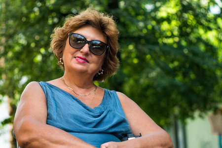 A mature lady in sun glasses. A happy middle-aged woman is smiling while sitting in the park on a sunny day. An outdoor horizontal shot.の写真素材