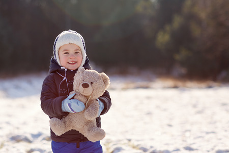 Portrait of a sweet little boy holding his teddy bear in a sunlit snowy fieldの写真素材