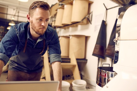 Handsome male entrepreneur in his clean and neat coffee roastery working with his laptop and a modern roasting machineの写真素材
