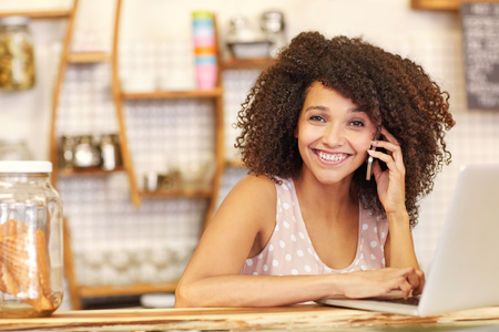 A young woman working on her laptop while speaking on the phone in a coffee shopの写真素材