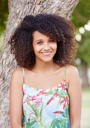 Beautiful mixed race woman happily smiling at the camera while leaning against a tree trunk in a parkの写真素材