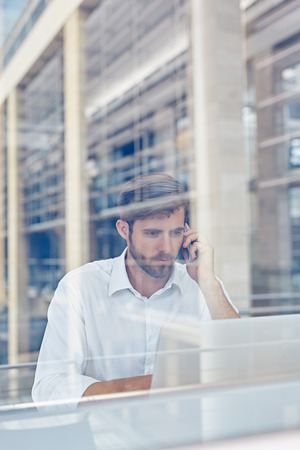 View through a window of a corporate executive working on his laptop and talking on a mobile phone