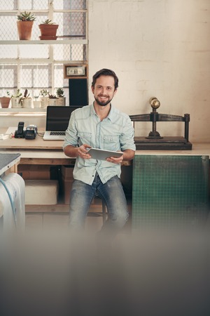 Portrait of a businessman in his design studio smiling at the camera while working on his digital tablet, sitting on a high stool in front of his workbenchの写真素材