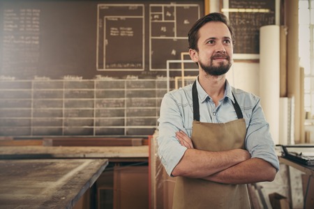 Cratsman looking away in a thoughtful manner with his arms folded while standing in his carpentry workshop and smiling to himself