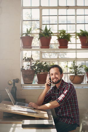 Smiling entrepreneur sitting at the desk of his studio office and looking pleasantly at the camera while listening to his cell phoneの写真素材