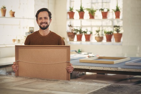Portrait of a young male professional framer holding a newly framed work while smiling confidently...