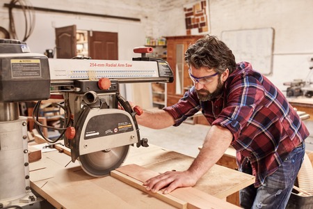 Skilled carpenter in his woodwork workshop, cutting a piece of wood with a radial arm circular saw,...
