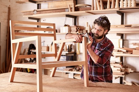 Serious furniture designer carefully sanding a chair frame that he is busy manufacturing in his woodwork studio, with shelves of wooden items behind himの写真素材