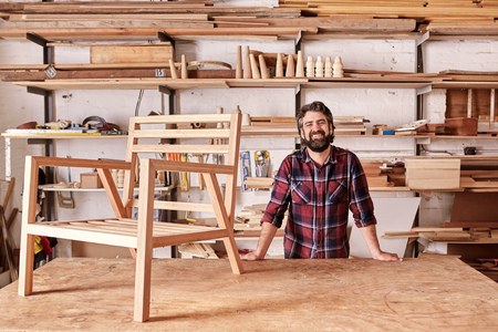 Portrait of a smiling craftsman with a rugged beard, resting on his workbench with a wooden chair frame on it, looking at the camera, with shelves of wooden planks and pieces behind himの写真素材